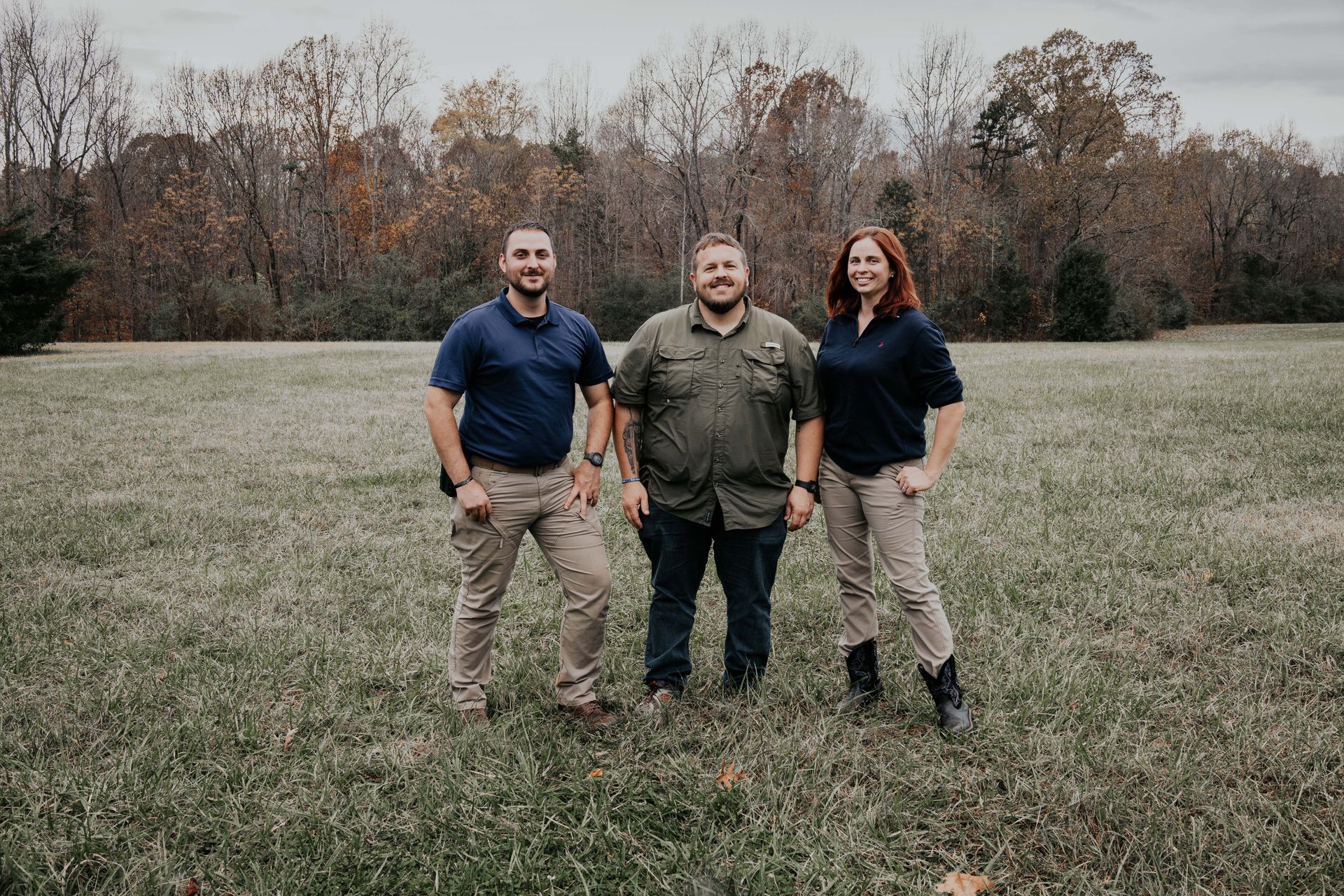 Three people standing in a field. Trees in the background. Man in center wearing olive shirt smiles. Others in blue shirts.