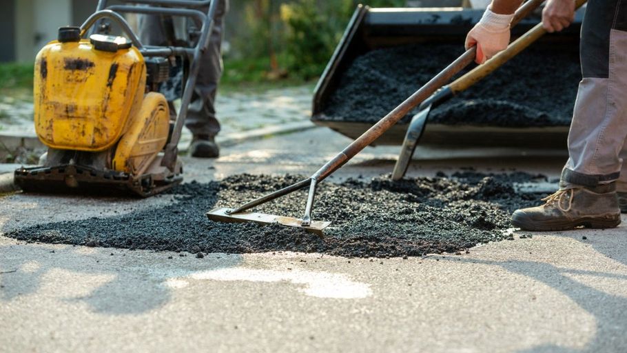 Workers spreading fresh asphalt with shovels beside a yellow plate compactor on a road repair site