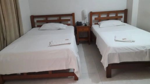 Dos camas individuales con sábanas blancas en una habitación sencilla, separadas por una pequeña mesita de noche.