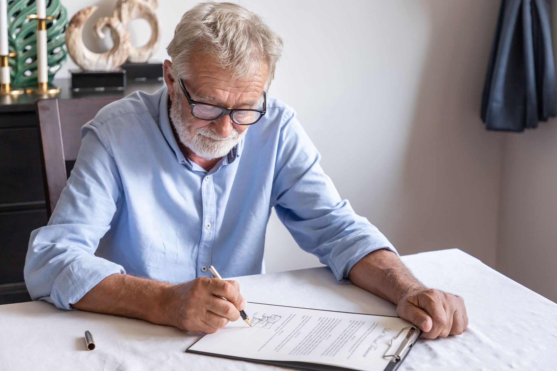 A senior man in a blue shirt and glasses signing a legal document at a table with a pen A senior man in a blue shirt and glasses signing a legal document at a table with a pen