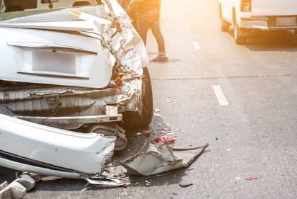White car heavily damaged in a road accident; debris scattered on asphalt, person standing nearby.