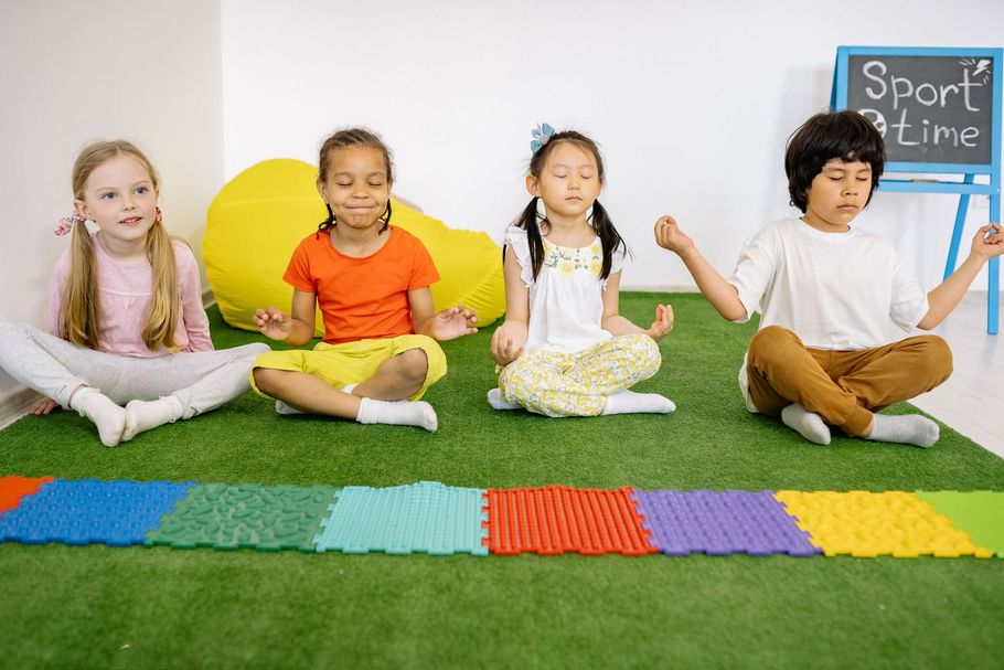 Four children sit cross-legged on mats in a classroom, holding hands during a yoga or meditation activity.
