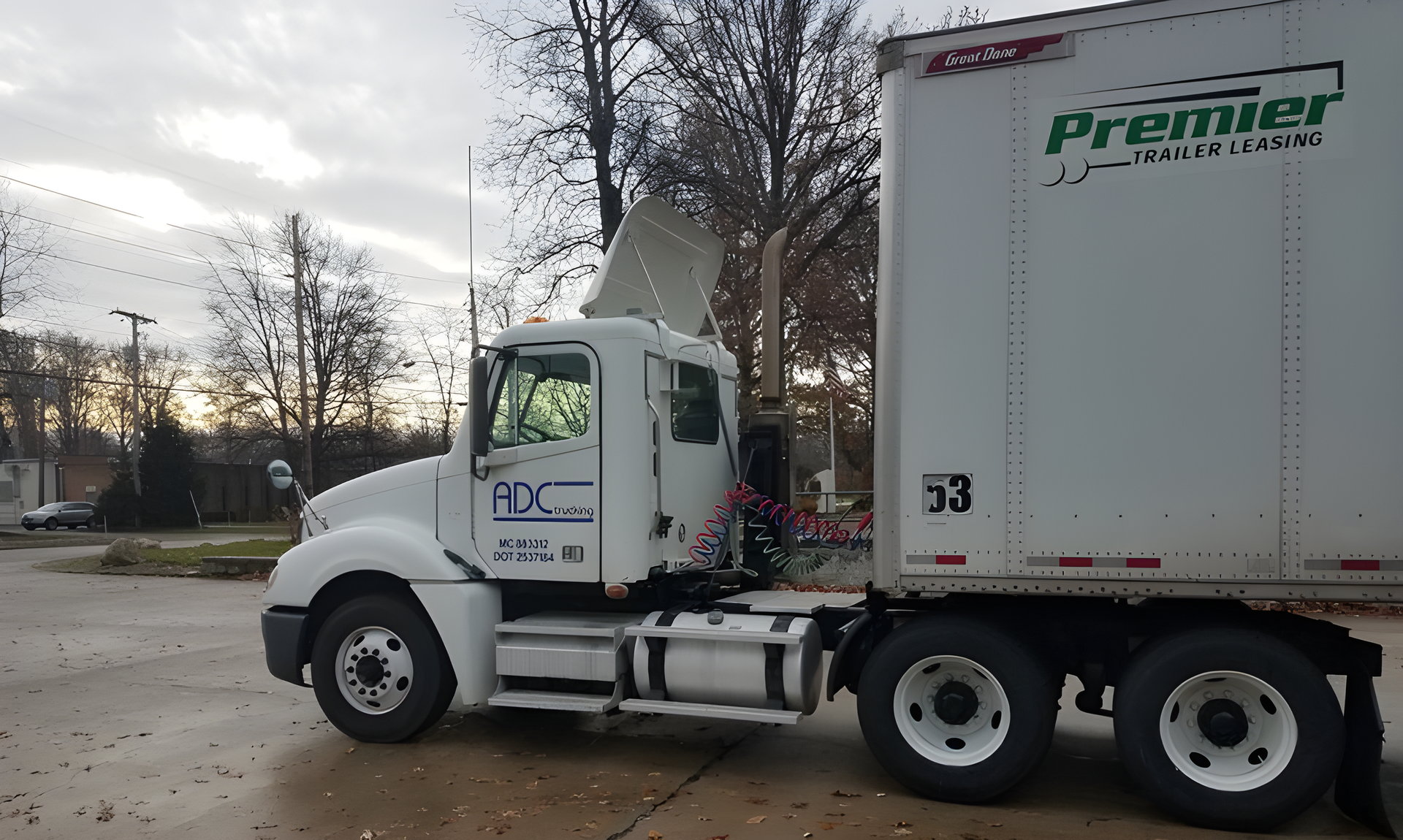 White Premier Logistics semi truck parked on a street beside leafless trees on an overcast day