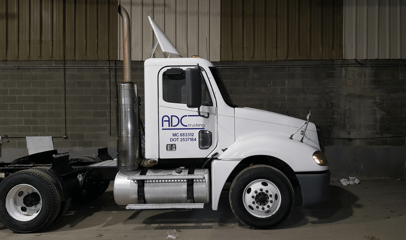 White semi-truck cab parked indoors against a concrete wall, viewed from the side.