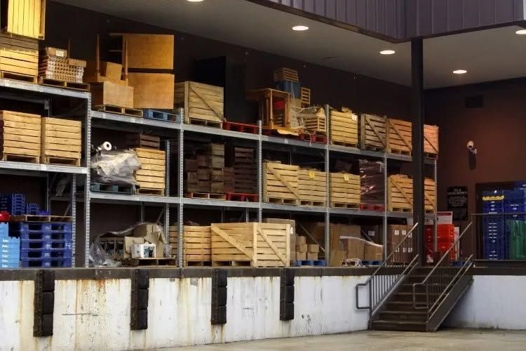 Warehouse interior with stacked wooden crates and pallets on metal shelves, with stairs at right