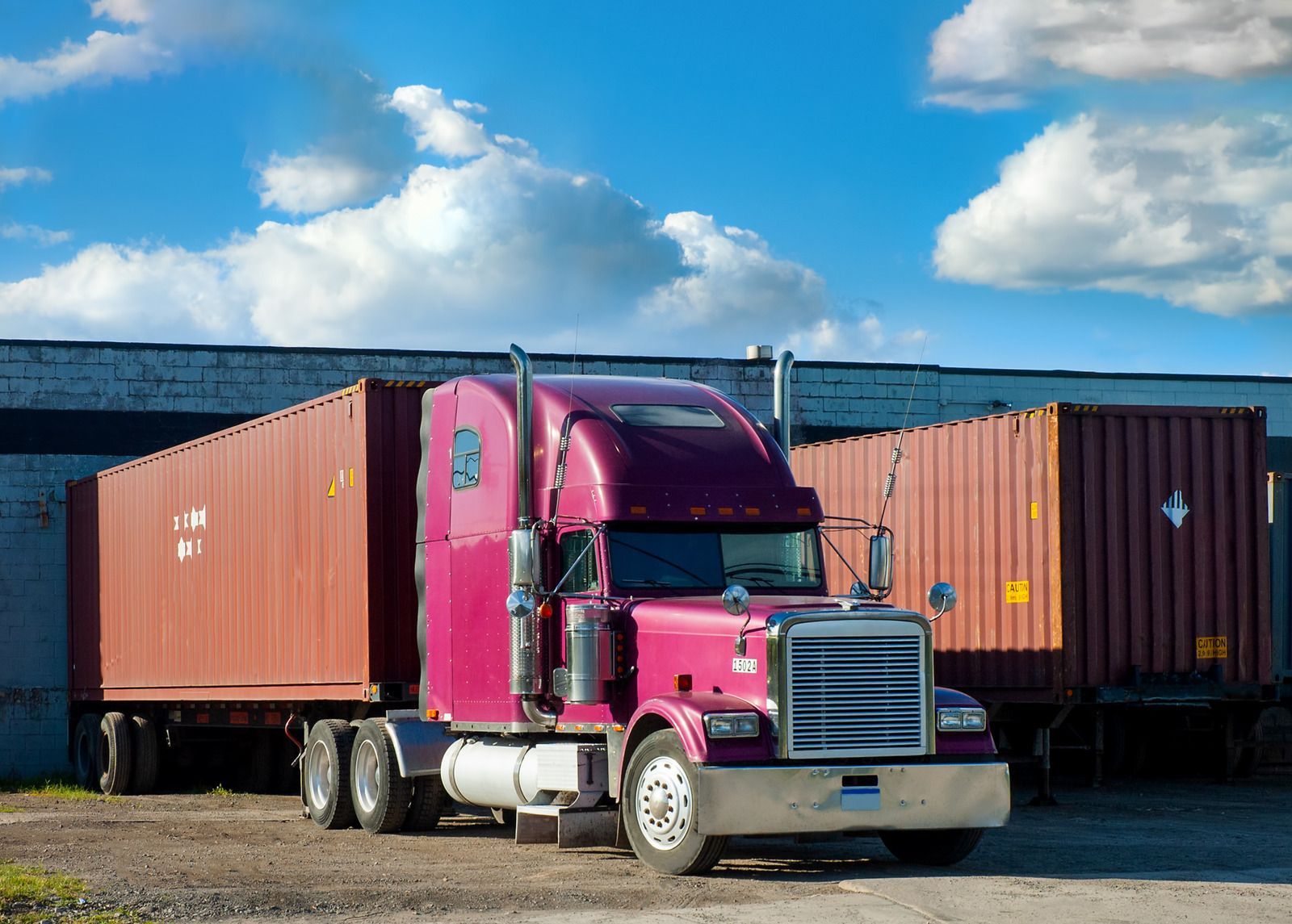 Pink semi-truck with shipping containers parked outside a warehouse under a blue sky