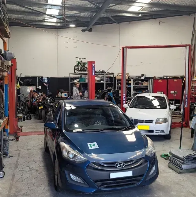 Inside a car repair shop: blue car in front, white car behind, mechanics and tools visible  — ATD Automotive in North Wollongong, NSW