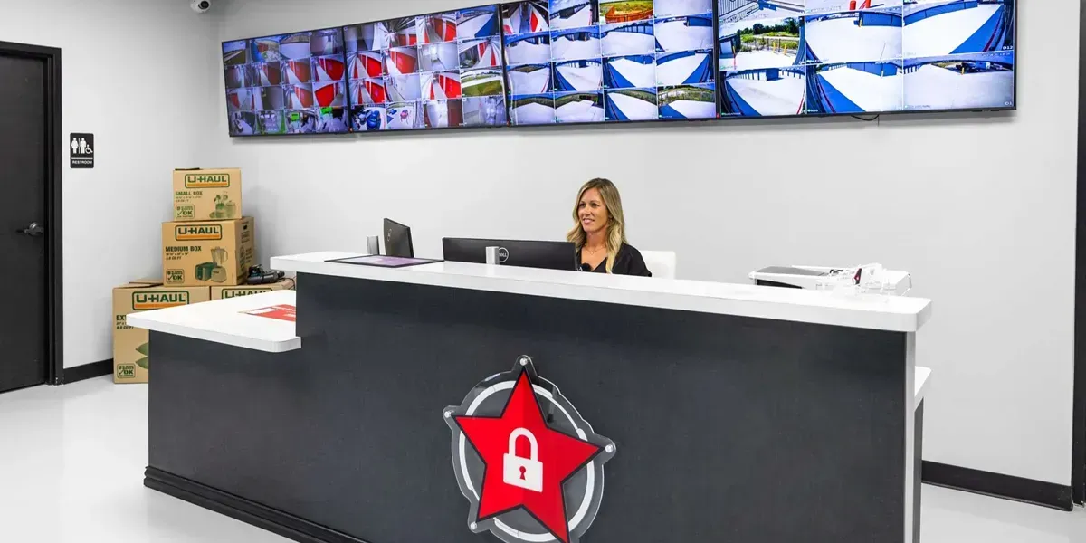 A person sitting at a reception desk with a security-themed star logo, in front of a wall with multiple security monitors.