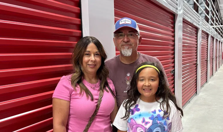 A family of three stands smiling in front of a row of closed, bright red storage unit doors.