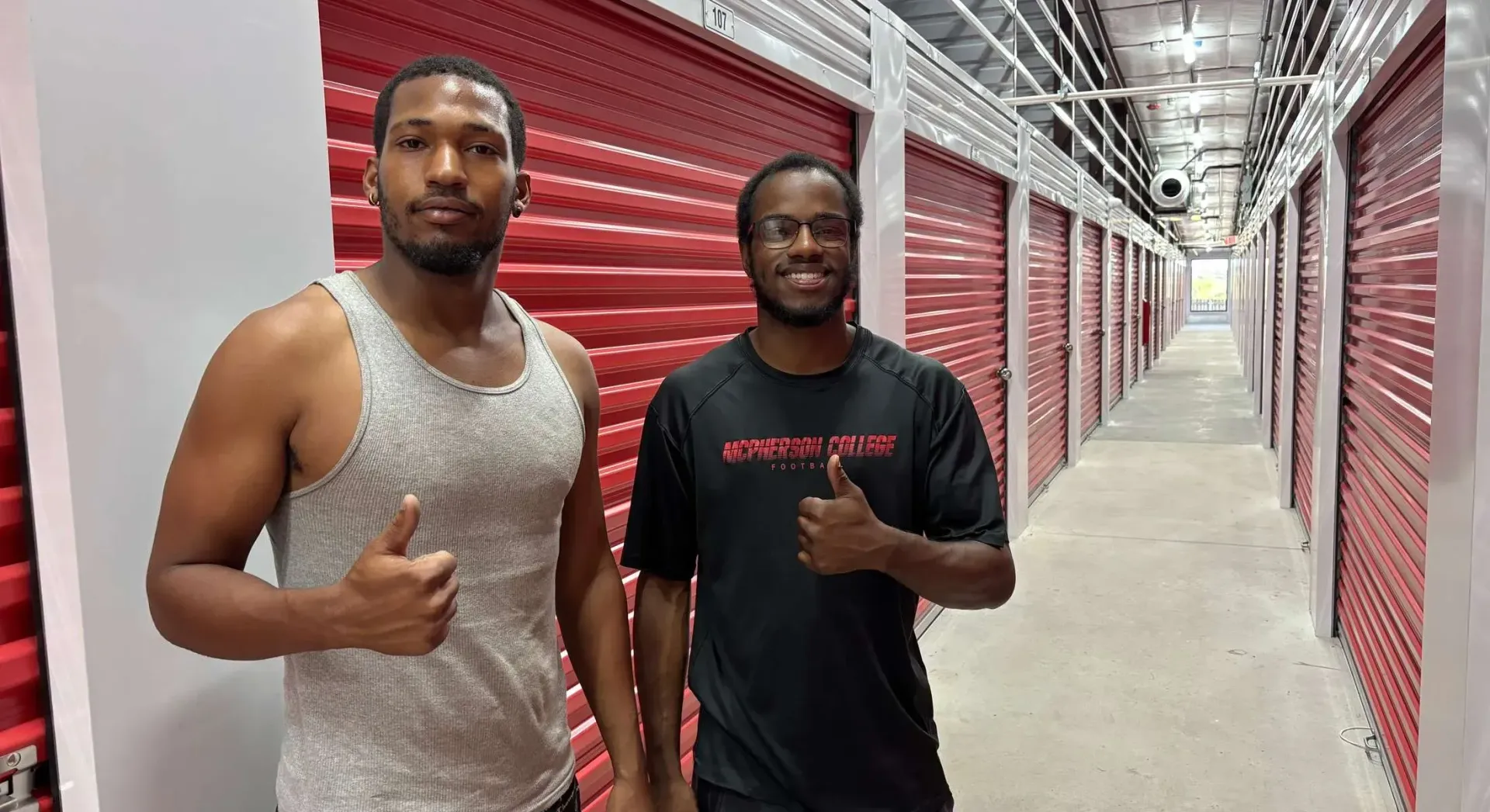 Two people wearing casual clothing stand in a hallway lined with red storage unit doors, giving a thumbs-up.