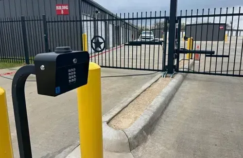 A keypad entry system mounted on a pole in front of a closed metal gate at a storage facility entrance.