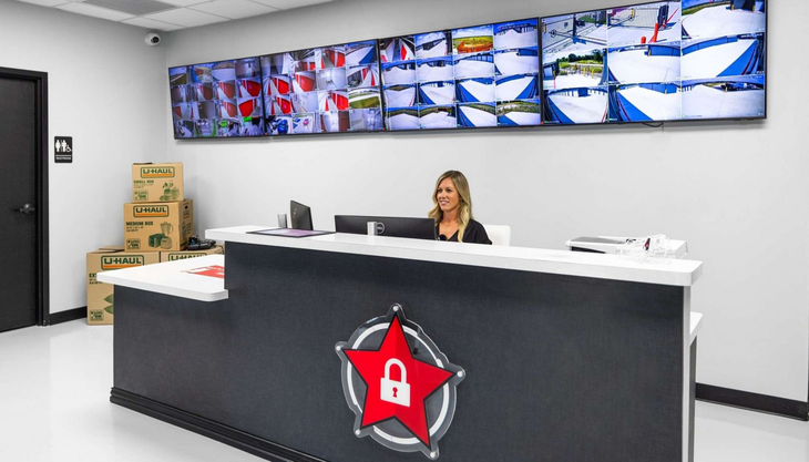 A staff member sits at a modern reception desk with a security logo, in front of a wall of surveillance monitors.