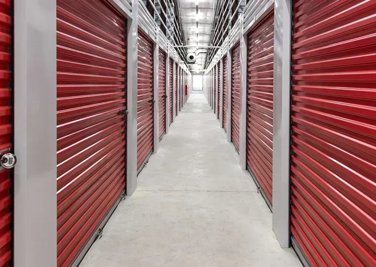 A long, brightly lit hallway in a self-storage facility lined with rows of red metal roll-up doors on both sides.