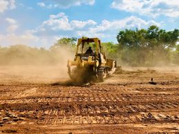 Yellow bulldozer grading a dusty dirt field with trees and blue sky in the background