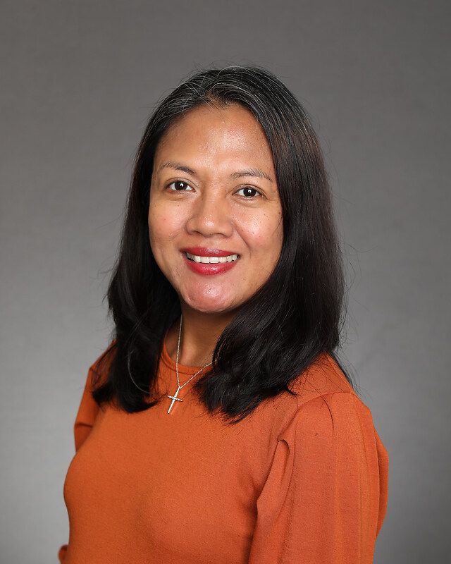 Woman with dark hair smiles, wearing an orange sweater, and a cross necklace. Gray background.