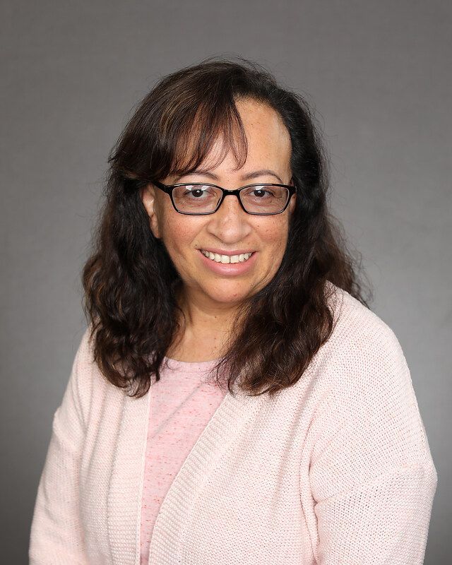 A woman wearing glasses and a pink sweater is smiling for the camera.