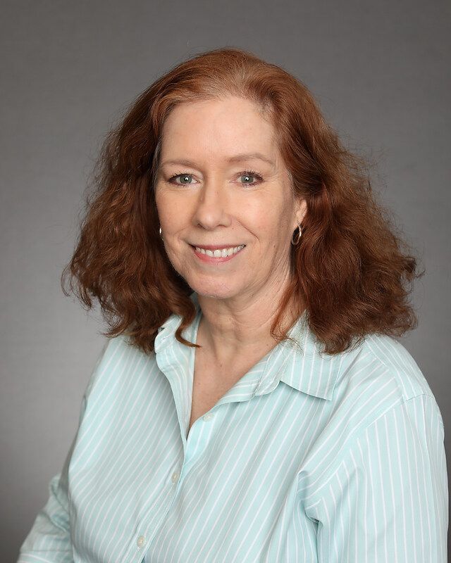 A woman with red hair is wearing a blue striped shirt and smiling for the camera.