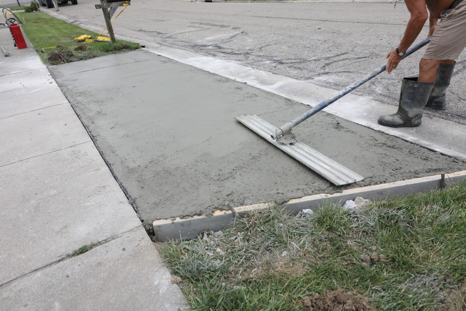A person levels wet concrete sidewalk with a long screed. They wear boots and shorts. The setting is outdoors.