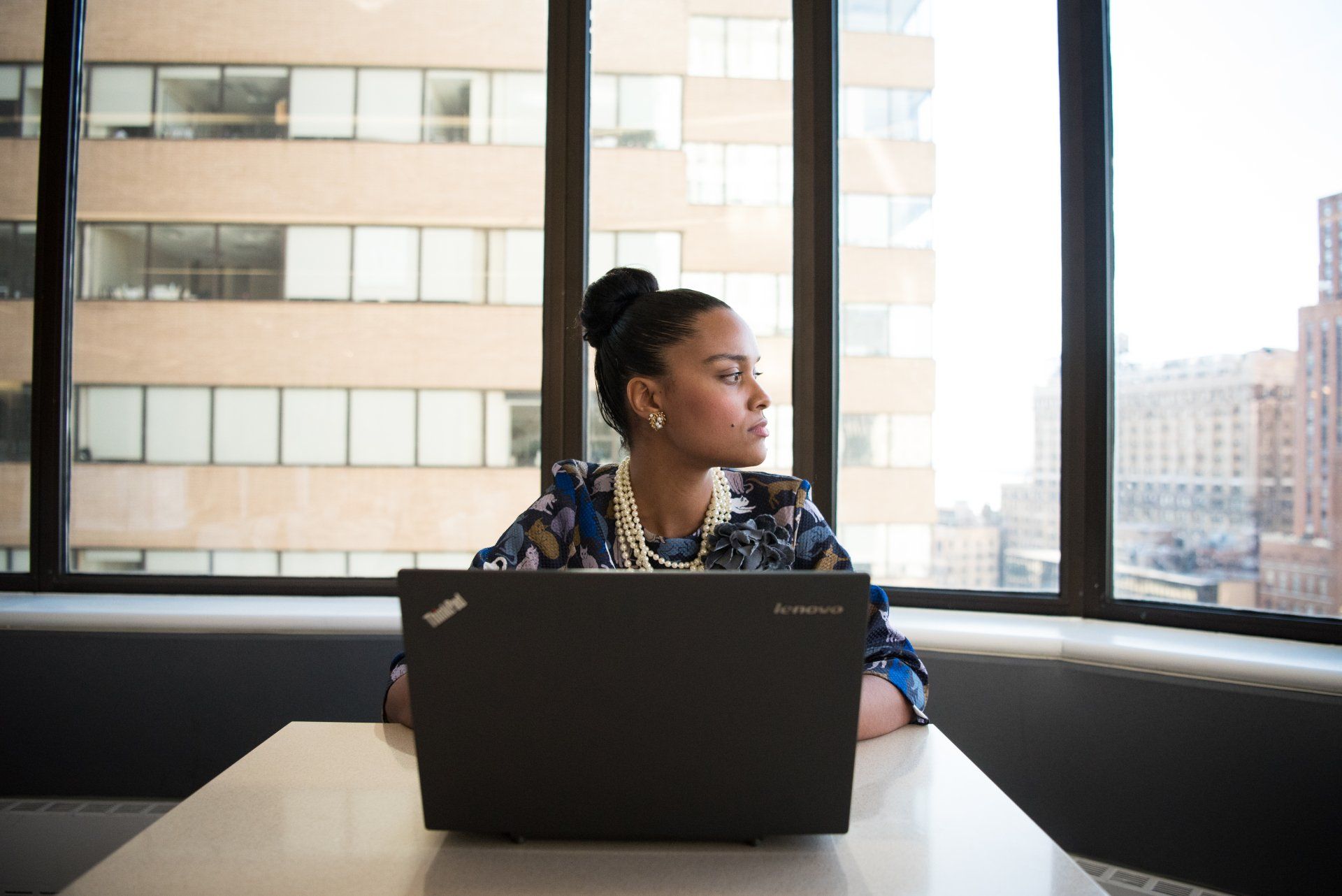 Person working on a laptop by a large window in an office, looking out thoughtfully