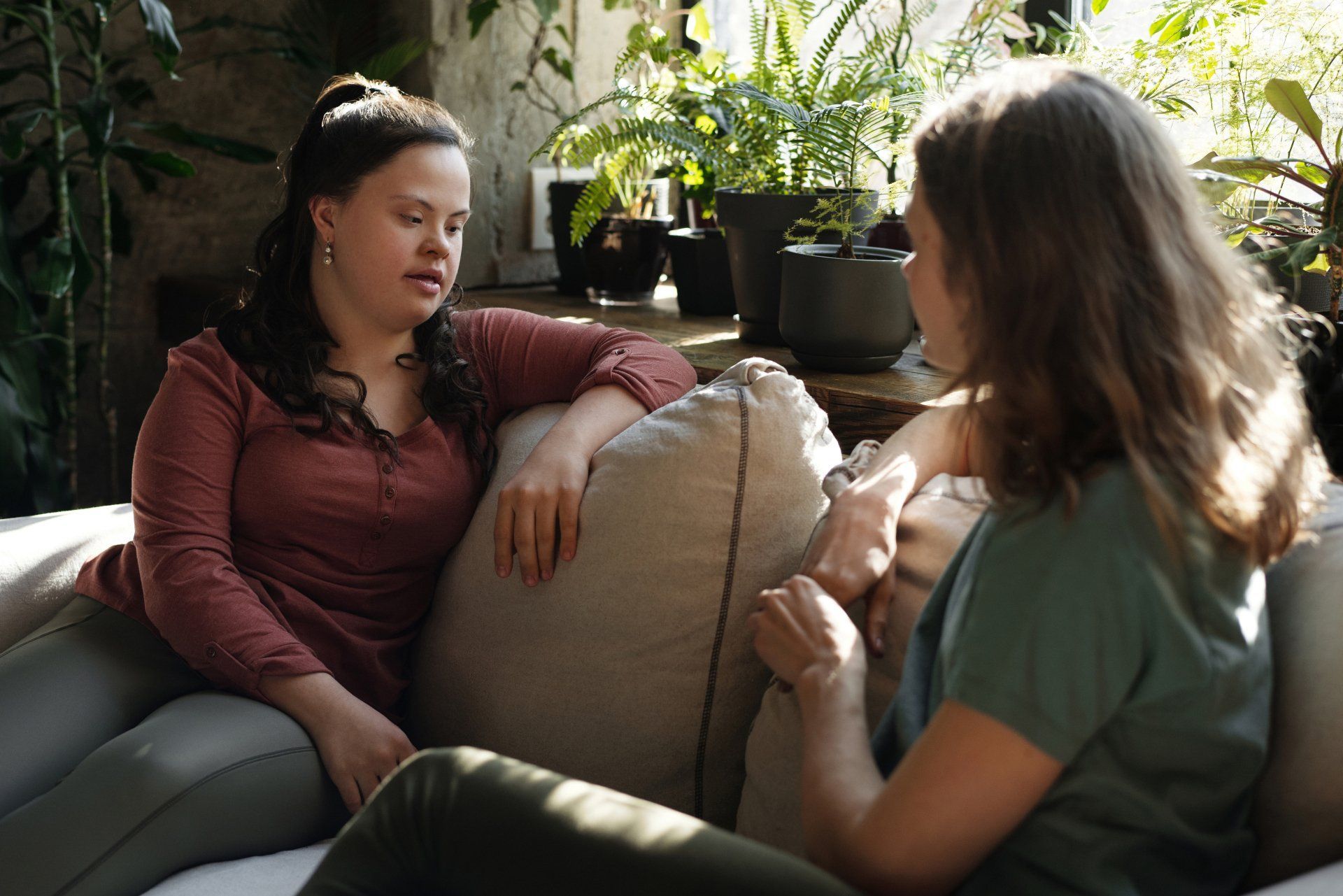 Two people talking on a couch in a sunlit living room with potted plants.