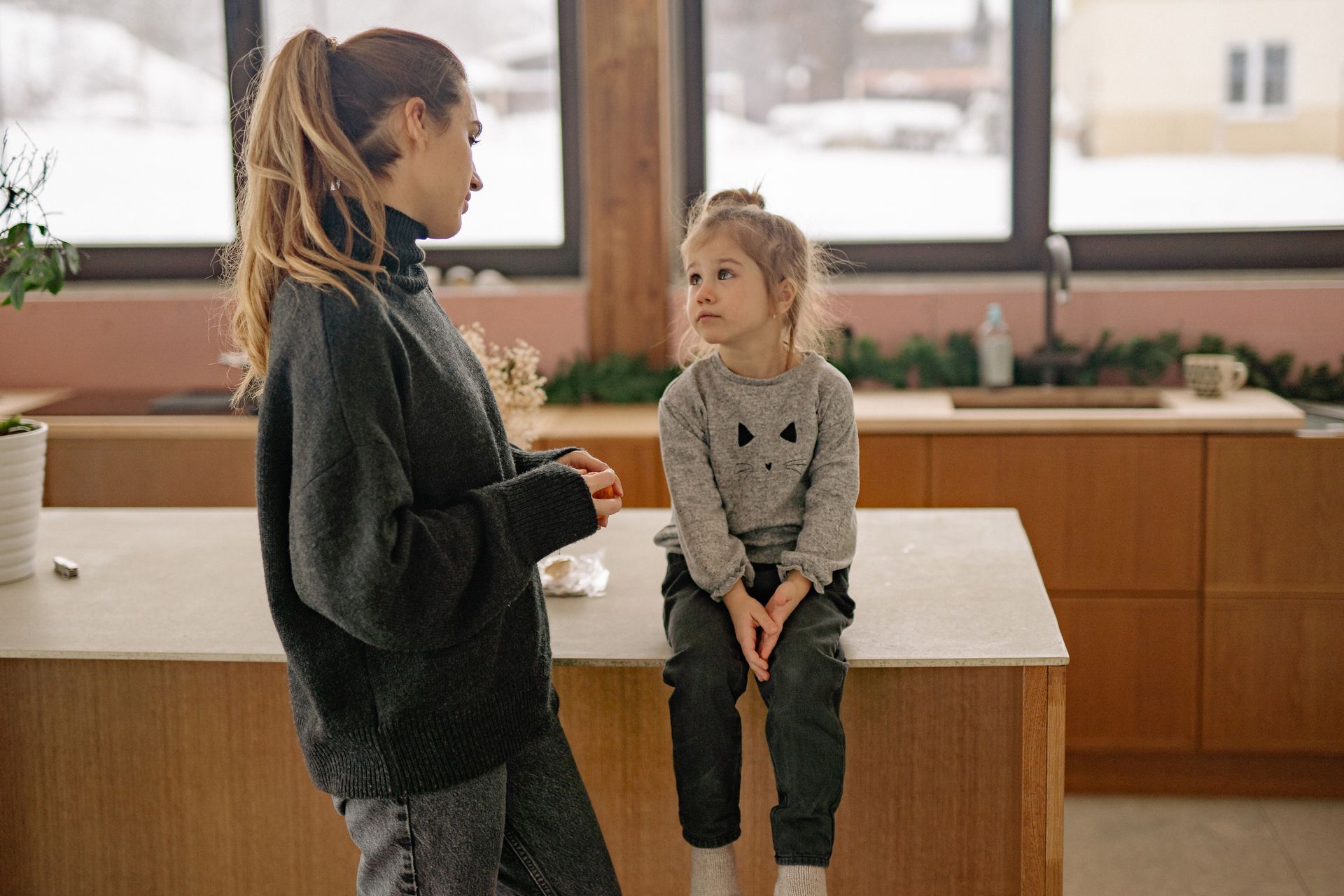 Two children talking in a bright kitchen, one standing and one sitting on a counter.