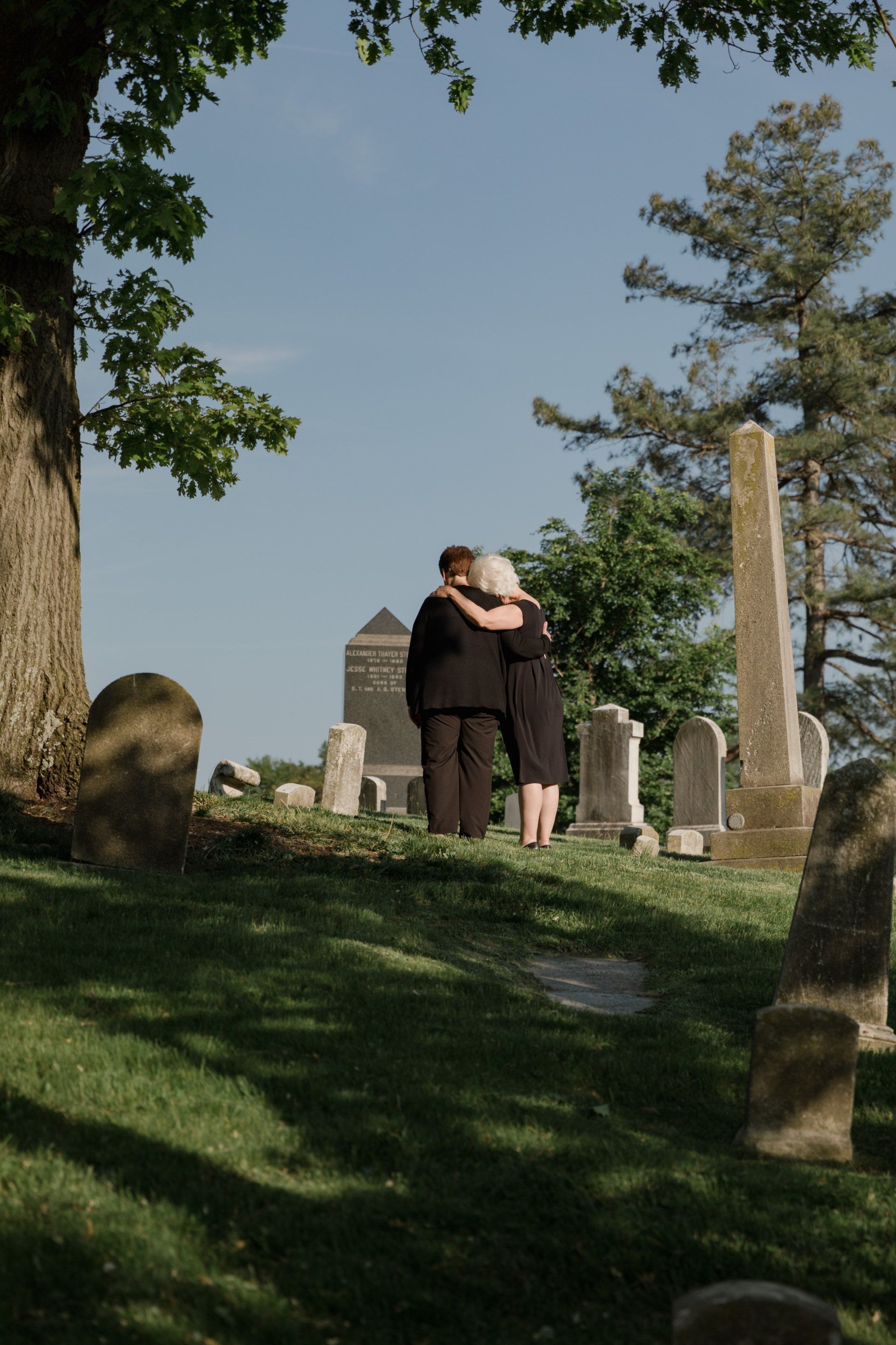 Two people embracing in a cemetery under trees, with gravestones and blue sky in the background.
