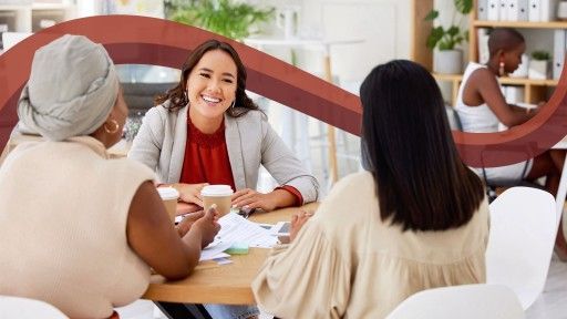 Three women in a meeting around a table, one smiling, and another woman working in the background.