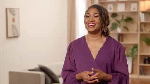 Woman in purple top smiles, hands clasped, in a living room with a couch and bookcase.