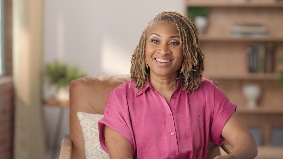 Woman with graying blonde locs smiles, wearing a pink shirt, seated in a brown chair, indoors.