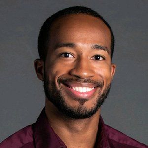 Man with dark skin smiling, wearing a burgundy shirt, against a gray backdrop.