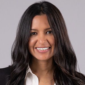 Woman with dark hair smiles, wearing a black blazer and white shirt.