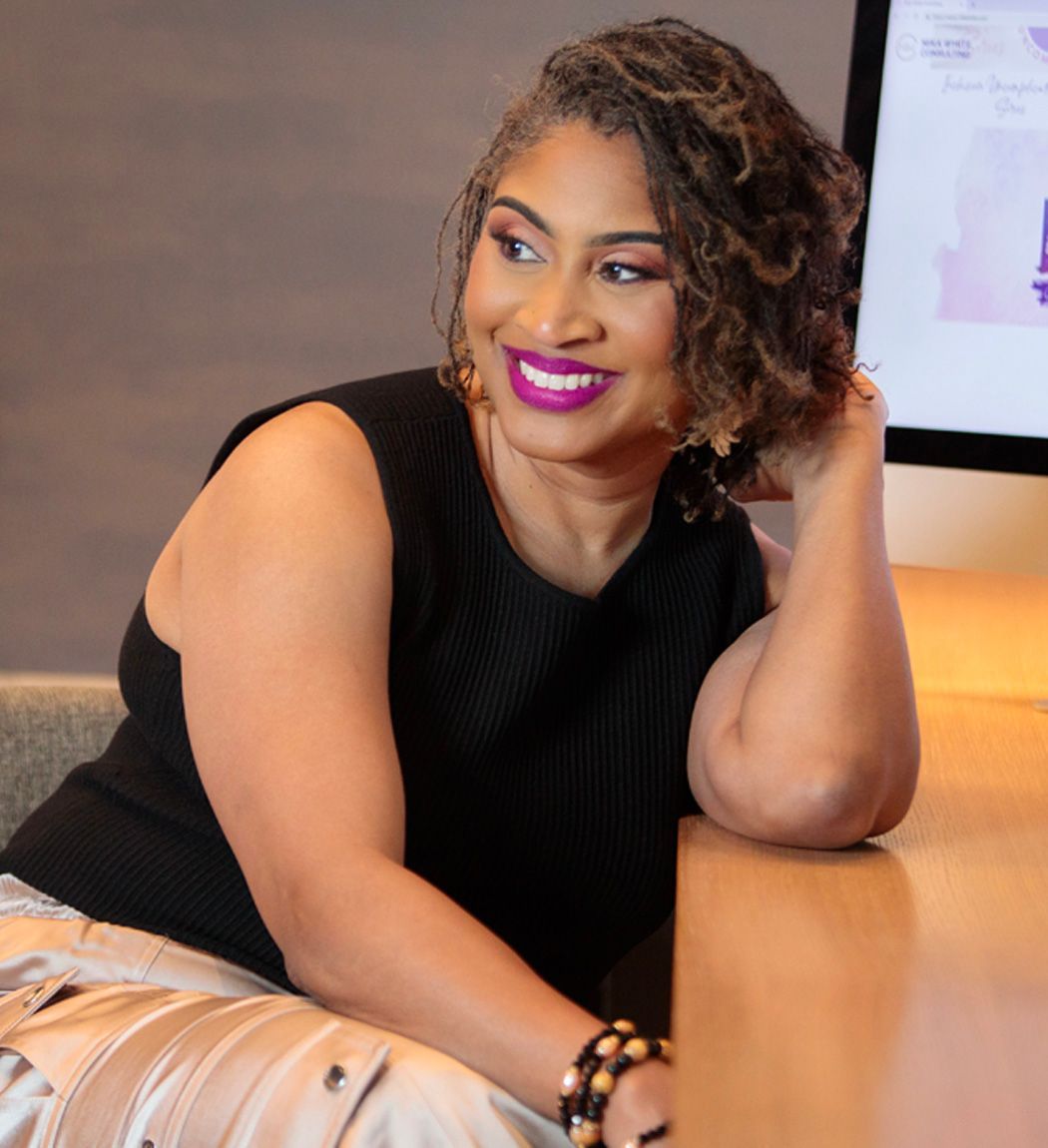Woman in black top, smiling, leaning on arm at a desk.