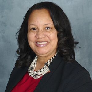 Woman smiling, wearing a red shirt, black blazer, and pearl necklace.