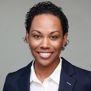 Woman in a blue blazer and white shirt, smiling, with short, dark curly hair, against a gray background.