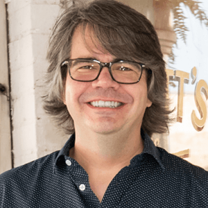 Man with glasses smiling, graying hair, blue patterned shirt; outdoors, bright setting.