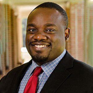 Man in a suit smiles at the camera, red tie, blue plaid shirt, blurred building background.