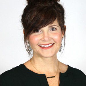 Woman with brown hair in a bun smiling, wearing a black shirt, earrings, and necklace.