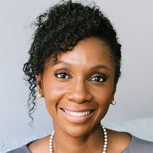 Woman with curly dark hair smiles, wearing pearl earrings and a necklace, in a light-colored setting.