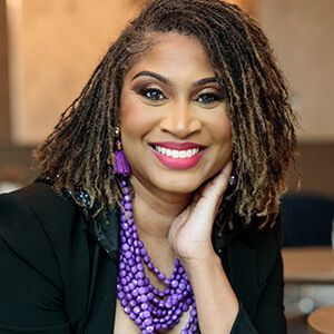 Woman with locs, smiling, wearing purple jewelry, black blazer.