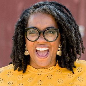 Woman with locs, black glasses, and yellow shirt smiles widely, showing teeth; gold earrings.