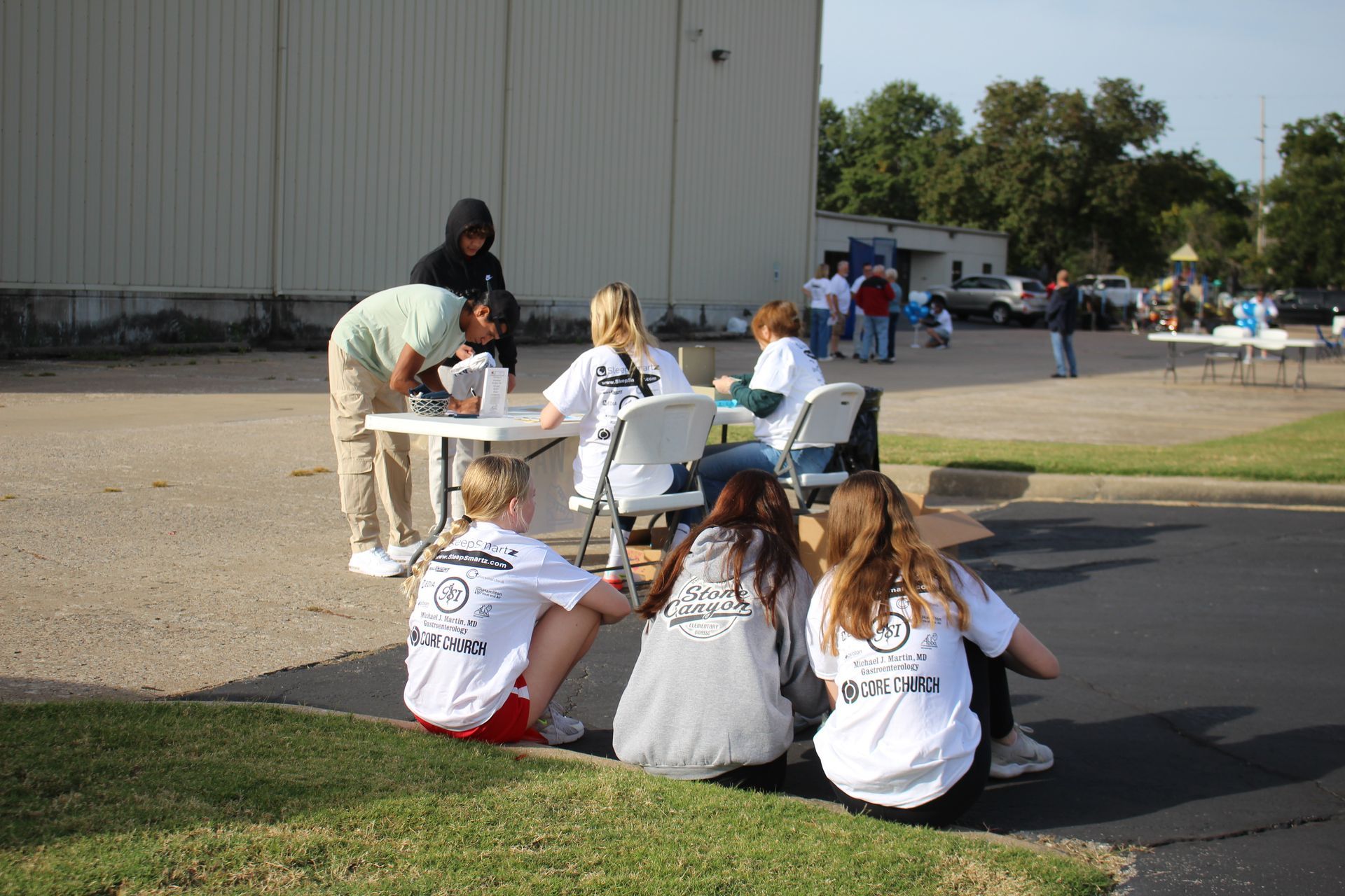A group of people are sitting around a table in a parking lot.