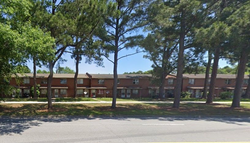 Brick apartment buildings behind tall pine trees along a street on a sunny day