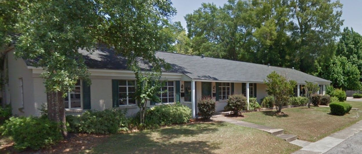 Single-story suburban house with gray roof, front porch, and landscaped yard under trees