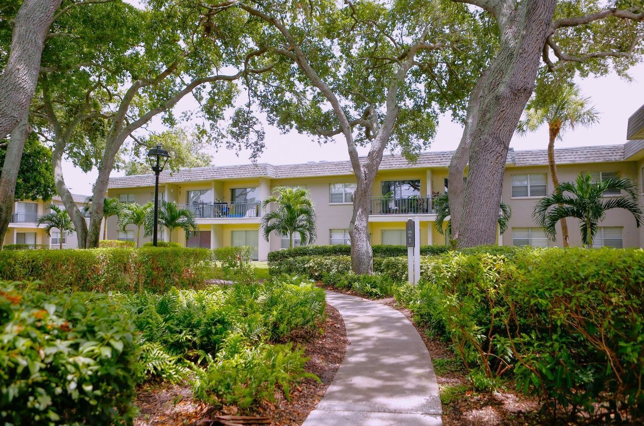 Curved garden path leading to a yellow apartment building surrounded by trees and shrubs.