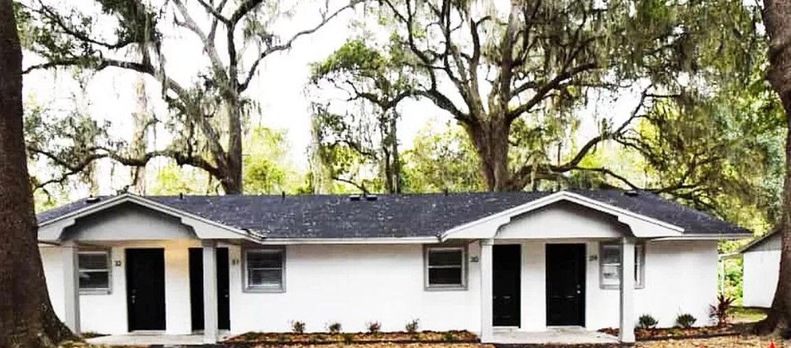 Small white house with black roof and front porch, surrounded by large trees and greenery.