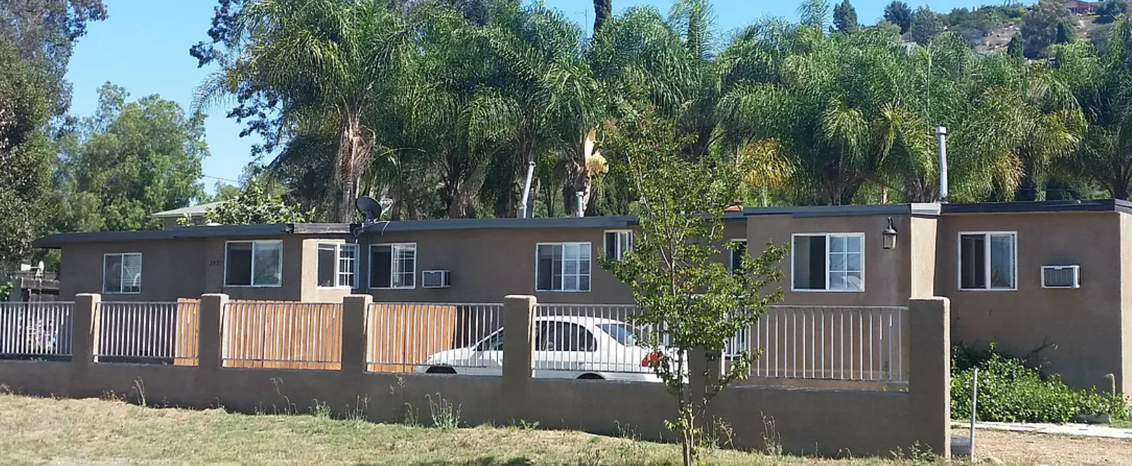 Low-rise apartment buildings with wooden fencing and parked cars along a tree-lined street