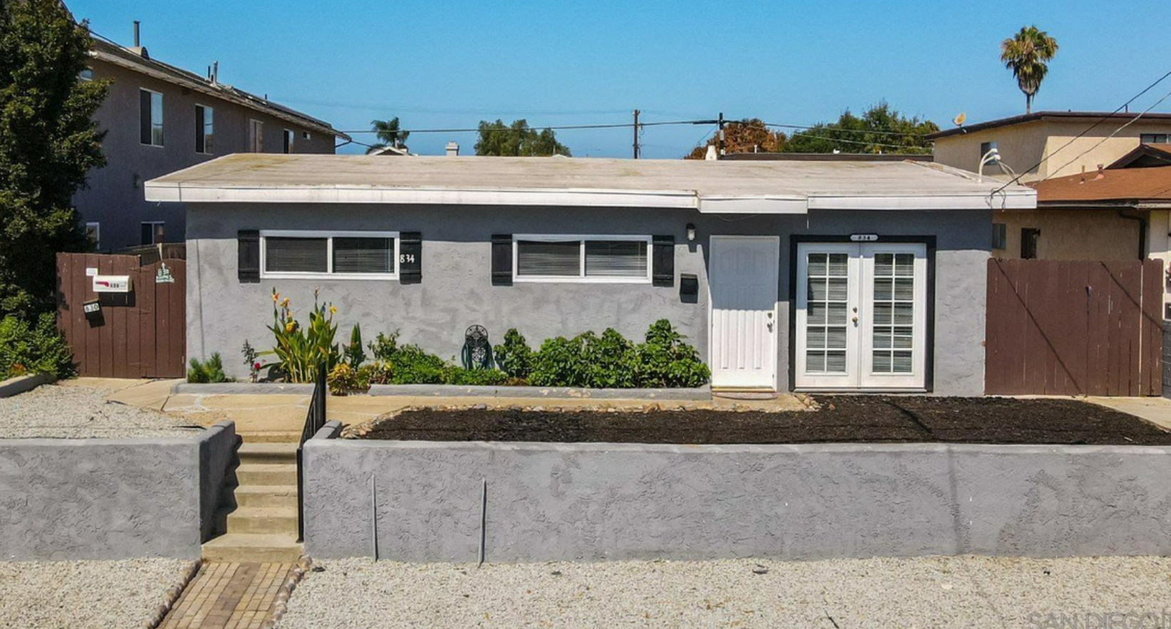 Single-story gray house with white trim, front steps, low wall, and a sunny residential street.