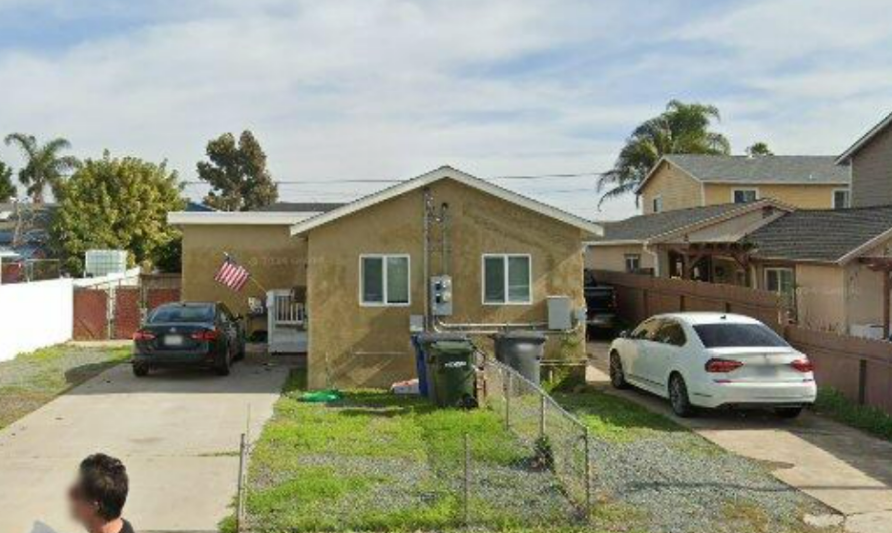 Single-story tan house with two parked cars, front fence, and a small American flag in a suburban neighborhood