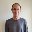Person in a gray shirt standing against a plain white wall, facing the camera with a slight smile.