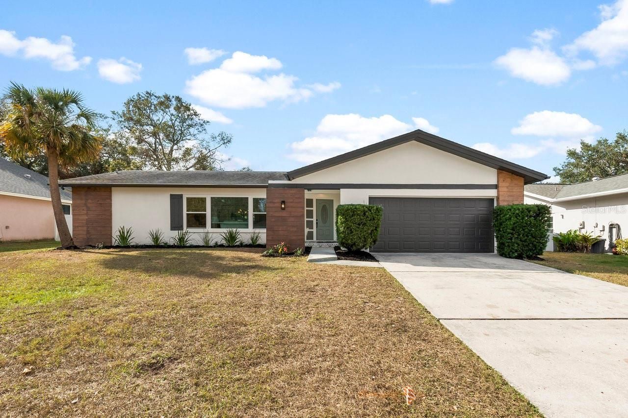 Single-story suburban house with a brick facade, gray garage, and a wide driveway under a blue sky.